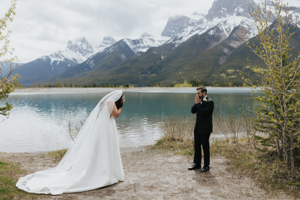 Groom covering his mouth with emotion during a first look with the bride beside a mountain lake in the Canadian Rockies
