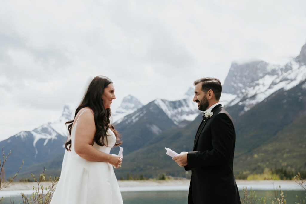 Bride and groom reading vows to each other beside a mountain lake with snow-capped peaks behind them