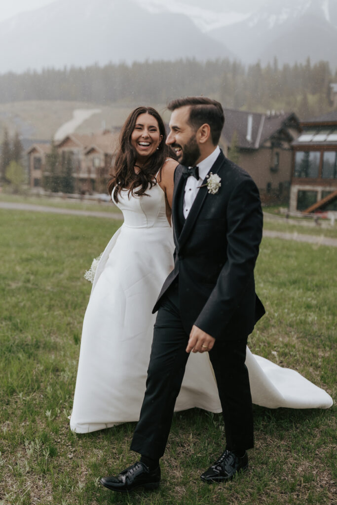 Bride and groom walking together through a grassy meadow with mountain homes and peaks in the background