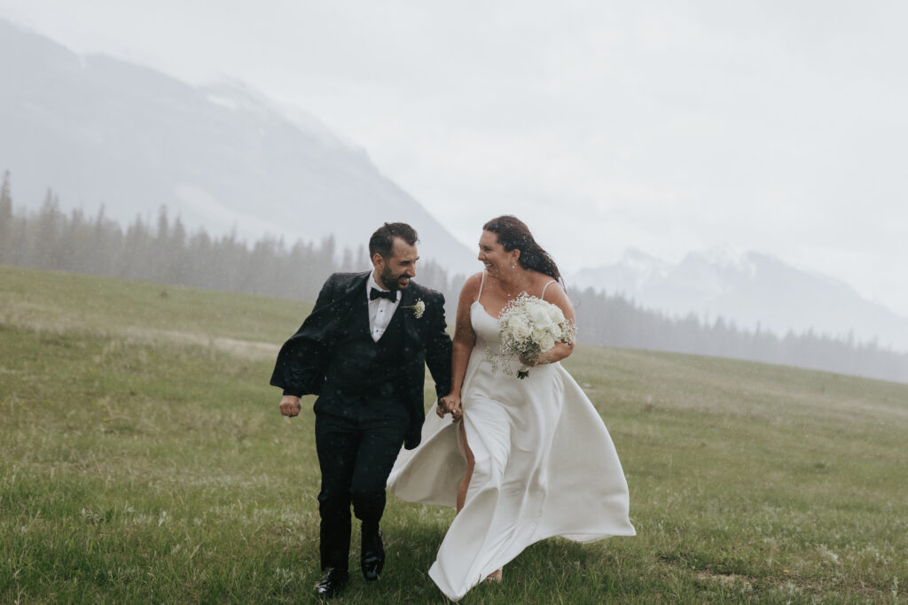 Bride and groom laughing together while walking through a mountain field during light rain
