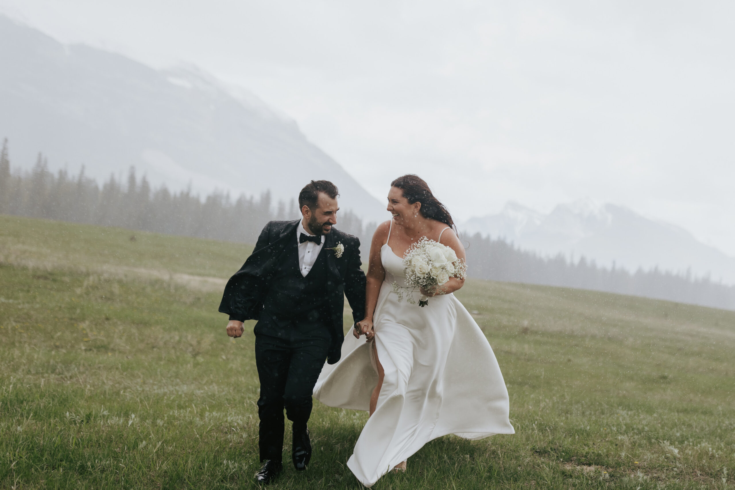 Bride and groom laughing together while walking through a mountain field during light rain