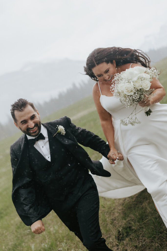 Bride and groom running hand in hand across an alpine meadow with misty mountains behind them