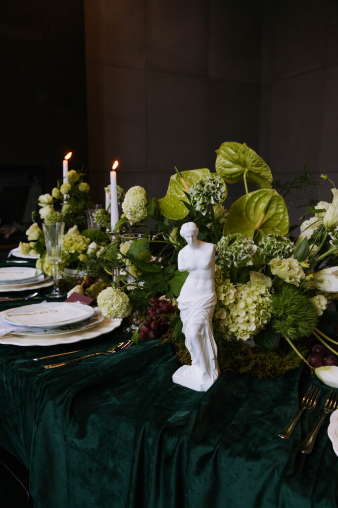 Long reception table styled with green florals, candles, grapes, moss accents, and classical sculpture decor
