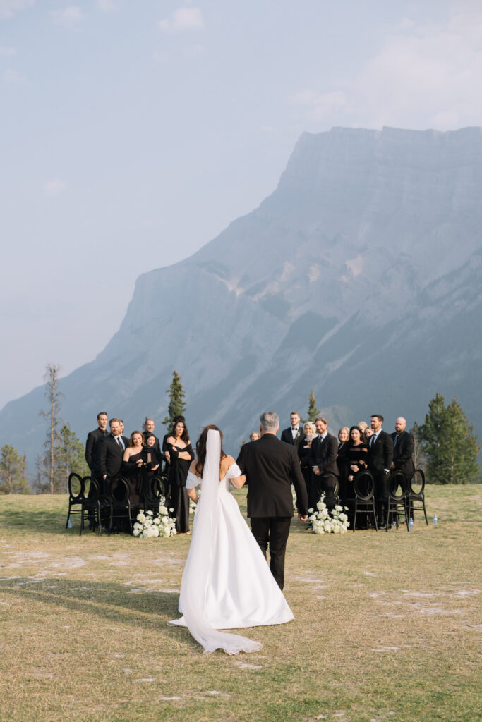 Bride walking down the aisle with her father during an outdoor ceremony with guests and mountains in the background