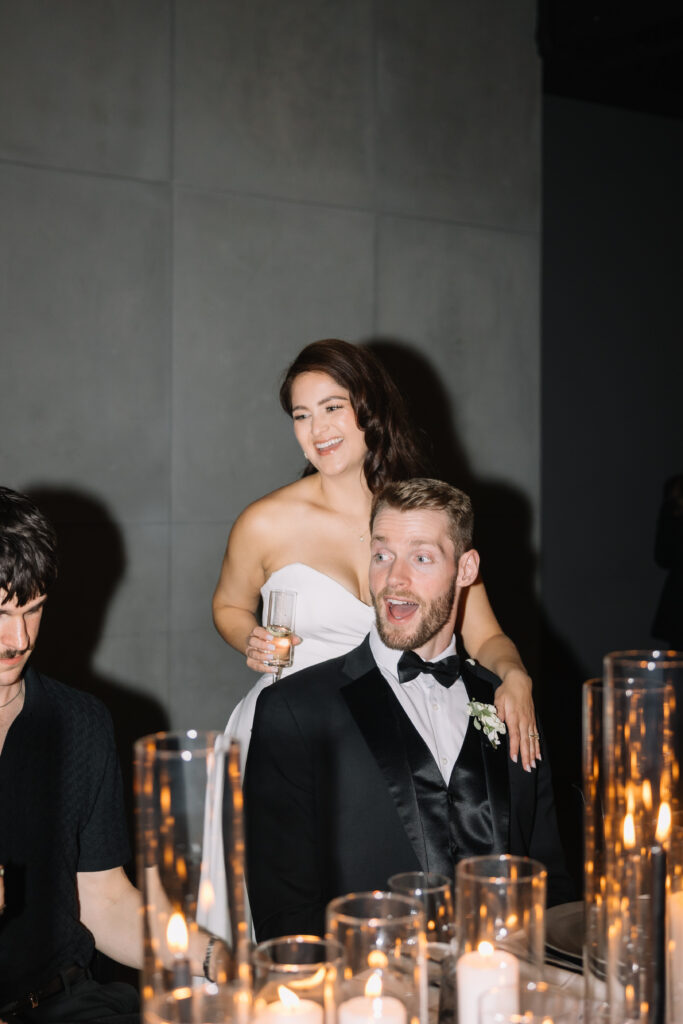 Bride standing behind groom while holding a champagne glass during a heartfelt reception speech at Mainspace